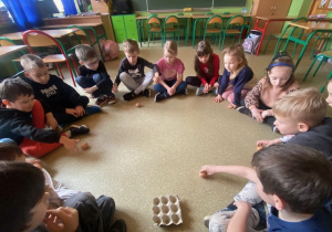 Uczniowie siedzą w kole i kręcą wylosowanymi jajkami na podłodze.