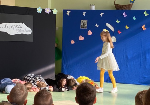 Uczennica w białej sukience kieruje się w stronę uczniów leżących na podłodze.