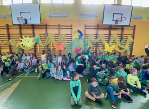 Uczniowie w zielonych strojach siedzą na podłodze w sali gimnastycznej.