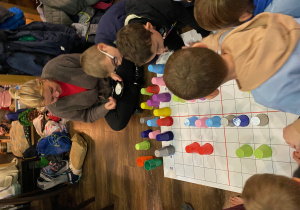 Uczniowie siedzą przy macie do kodowania i wykonują zadanie z użyciem kolorowych kubków.