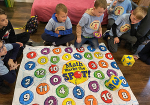 Uczniowie siedzą przy macie pokrytej kolorowymi cyframi i z użyciem kostek wykonują działania matematyczne.