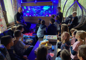 Uczniowie siedzą przed akwarium słonowodnym i słuchają opowieści o koralowcach.
