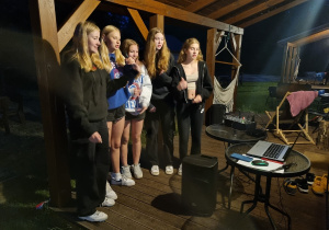 Grupa dziewcząt śpiewa piosenkę do mikrofonów czytając tekst z laptopa.
