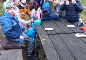 Uczniowie siedzą przy ławce i jedzą kiełbaskę.