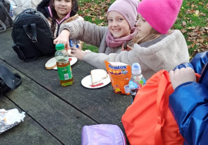 Uczniowie siedzą na dworze przy stole, piją i jedzą.