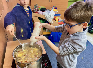 Jeden uczeń wlewa barszcz do garnka z zupą, a drugi miesza zupę.