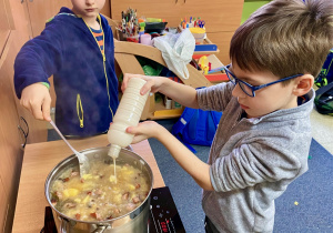 Jeden uczeń wlewa barszcz do garnka z zupą, a drugi miesza ową zupę.