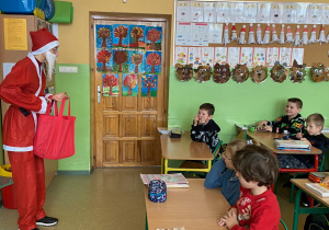 Mikołaj stoi z czerwoną torbą przed uczniami siedzącymi w ławkach.