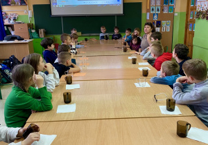 Dzieci siedzą przy długim stole, a przed nimi stoją na serwetkach szklanki.
