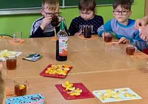 Dzieci piją herbatę, na stole na deskach ułożone są jabłka, imbir, cytryna, pomarańcza, obok leżą goździki, cynamon, stoi sok malinowy.