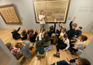 Uczniowie siedzą przed zdjęciem Łodzi i odpowiadają na pytania pani przewodnik.