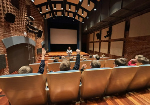 Uczniowie siedzą w sali kinowej przed prowadzącą warsztaty.