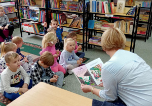 Przedszkolaki siedzą na dywanie i słuchaja bajki czytanej przez bibliotekarkę.
