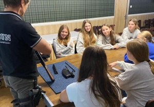 Grupa dziewcząt siędzi przy stole, na którym leżą elementy rozłożonego karabinu. Instruktor prezentuje czynność czyszczenia broni palnej.