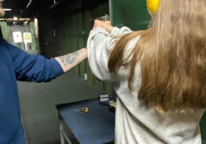 Nastolatka celuje do tarczy z pistoletu sportowego, instruktor poprawia ułożenie jej dłoni na rękojeści pistoletu.