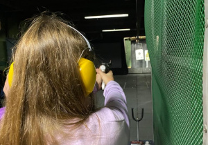Dziewczyna w filetowej bluzie mierzy do tarczy z pistoletu sportowego. Zdjęcie zrobione znam jej prawego ramienia, w oddali widać tarczę.