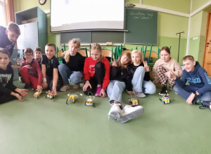 Grupa dzieci siedzi na podłodze sali lekcyjnej, przed nimi stoją zbudowane przez nich roboty z klocków LEGO.