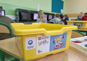 Na pierwszym planie żółte pudełko z etykietą LEGO Spike Prime, w tle dzieci pracujące przy komputerach.