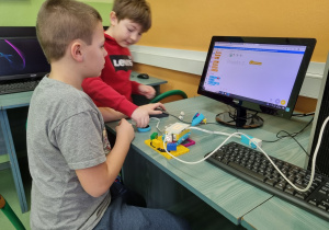 Dwójka chłopców pracuje przy komputerze, do którego kablem podłączony jest zestaw LEGO, na monitorze widać fragment skryptu.