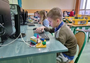 Rząd dzieci siedzących przy stanowiskach komputerowych. Na pierwszym planie chłopiec piszący na klawiaturze, obok stoi zestaw LEGO Spike Prime.