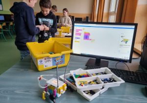 Na pierwszym planie zestaw LEGO Spike Prime w zółtym pudełku, obok tacki z klockami. Widać zbudowany robot oraz monitor, na którym wyświetlony jest gotowy skrypt.