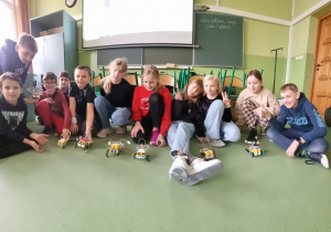 Grupa dzieci siedzi na podłodze sali lekcyjnej, przed nimi stoją zbudowane przez nich roboty z klocków LEGO.