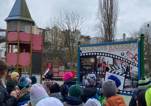 Uczniowie stoją przy plac zabaw, a w tle tablica informacyjna nt filmu Magiczne Krzesło
