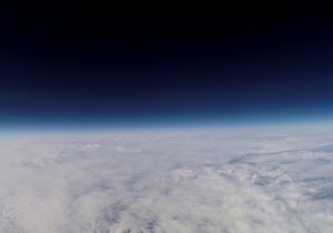 Zdjęcie pokazujące bardzo wysokie warstwy atmosfery. Niebo jest czarne, widać warstwę cienką błękitu a poniżej białe chmury.